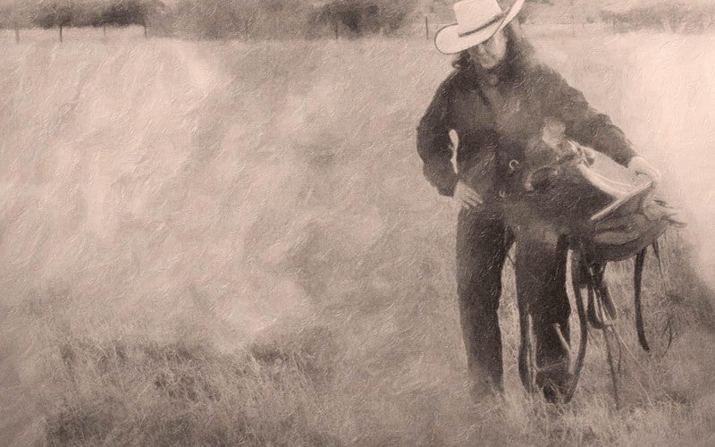 A muted image of Kathy in a grass field wearing a cowboy hat and holding a horse saddle.