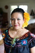 Maria de Jesus Gonzalez Laguna stands indoors. She wears a black outfit with multicolored embroidery, as well as a yellow flower in her hair.