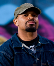 Michael "Mic" Crenshaw stands in front of a blurred purple and blue background. He wears a denim jacket, a black t-shirt, and a gray hat. 