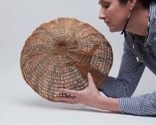 Sara Siestreem wearing a long sleeved blue shirt. She is leaning her arms on the ground and holding a woven basket in her hands. 