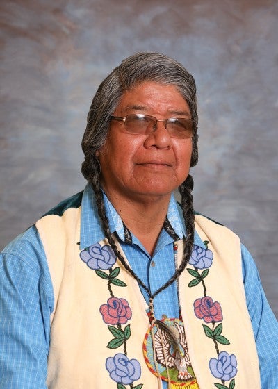 Portrait of Wilson Wewa against a gray background. He is wearing a cream embroidered vest and a blue collared shirt.