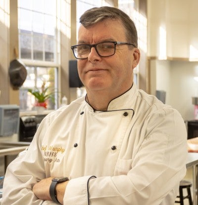 Frank Murphy stands in a kitchen with his arms folded wearing a white chef uniform.