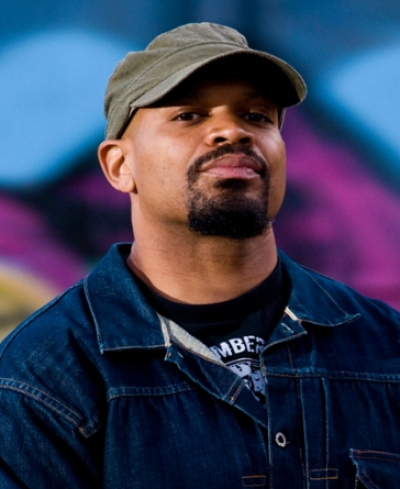 Michael "Mic" Crenshaw stands in front of a blurred purple and blue background. He wears a denim jacket, a black t-shirt, and a gray hat. 