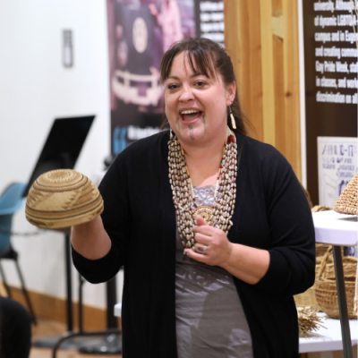 Stephanie Craig is standing in front of an audience holding a small woven basket. She is wearing a gray shirt with black cardigan and is gesturing with her left hand.