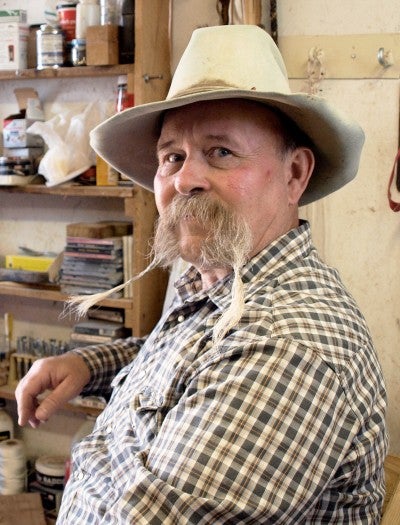 Dan Fowler sits next to a tall wooden shelf. He wears a plaid button-up shirt and a white cowboy hat.