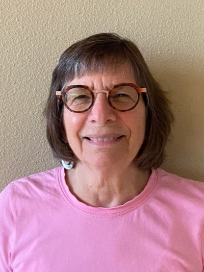 Irene Young wearing a pink shirt and standing against a tan wall.