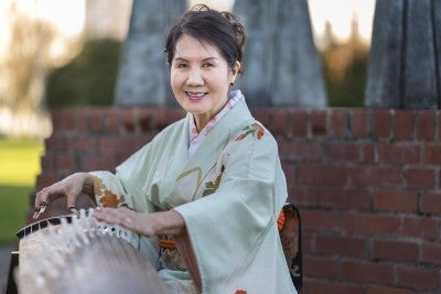 The artist, Masumi Timson, smiling as she plays the koto, a traditional Japanese stringed instrument.