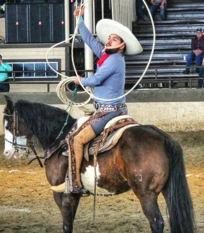 Josue Mendoza riding a horse and performing rope skills. He is wearing a white cowboy hat, blue shirt, and tan leather chaps.